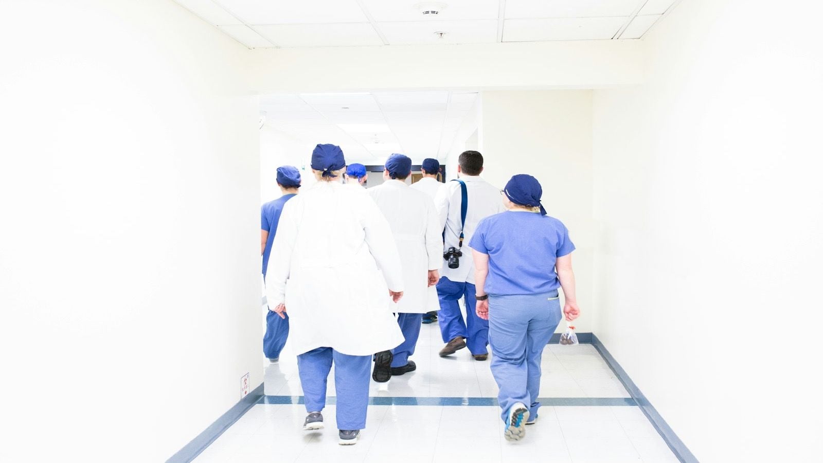 Care team of doctors and nurses in scrubs and white coats collaborating in a hospital hallway during a team huddle, the kind of internal meeting an AI meeting notetaker for healthcare teams transcribes